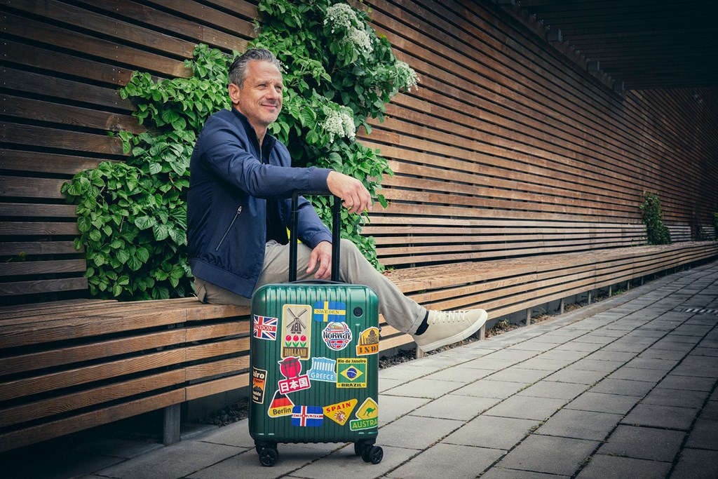 Leende man sitter på en bänk med en resväska täckt av reseklistermärken bredvid en trävägg.