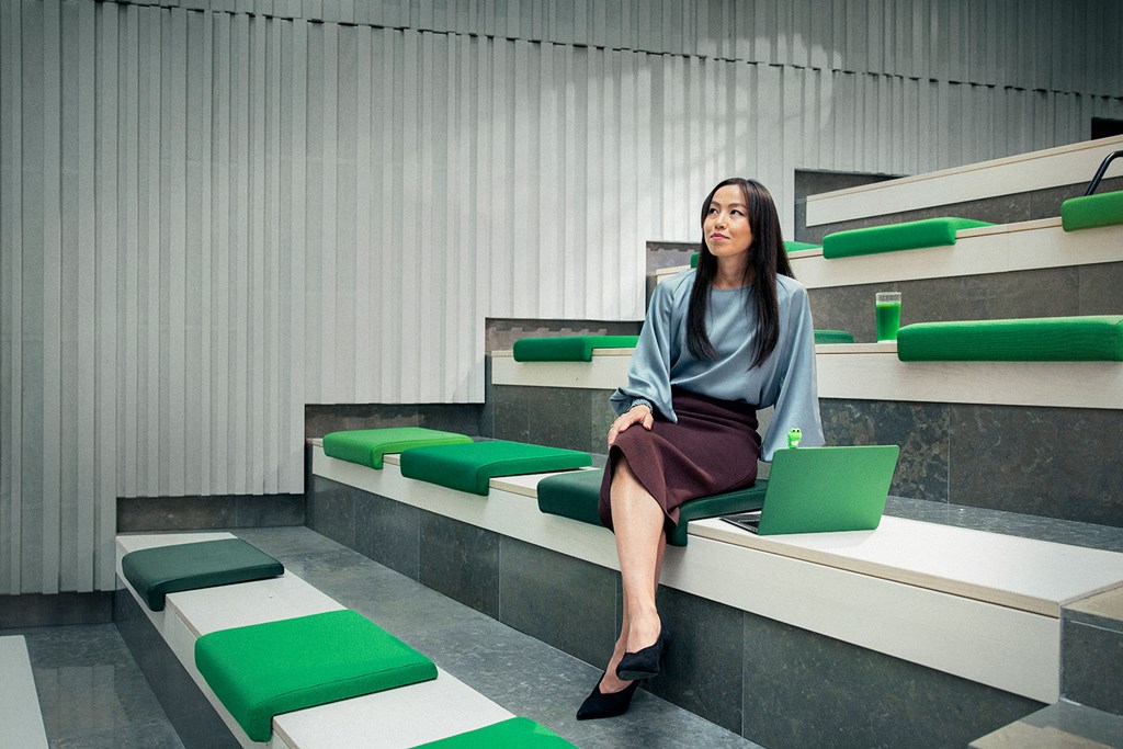 Professional woman seated in a modern room with green accents, laptop and drink beside her.