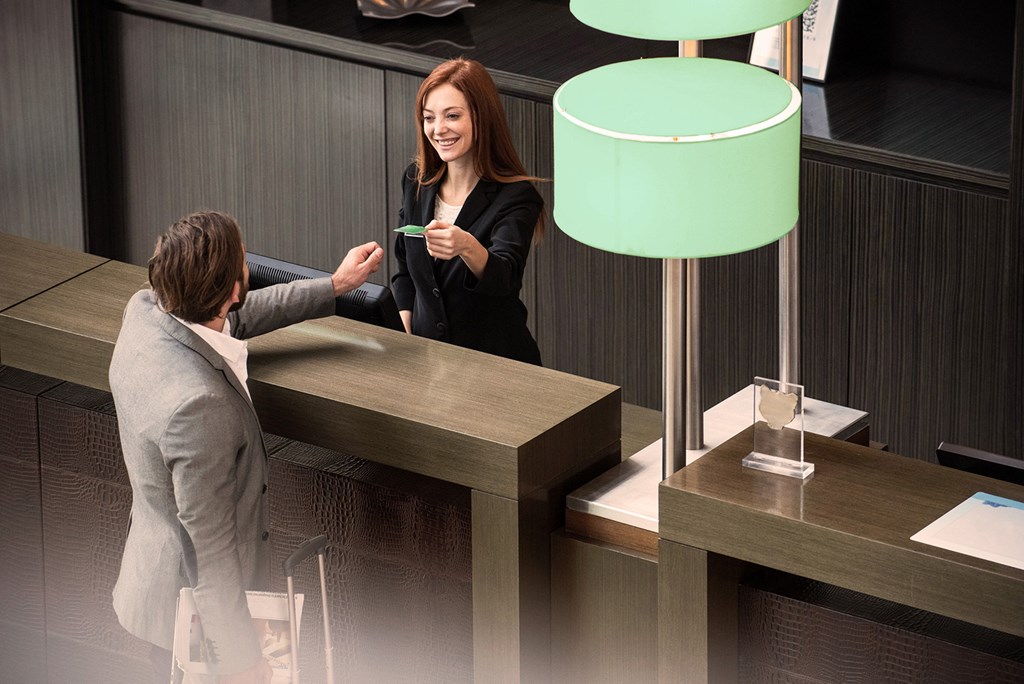 A woman behind a counter smiles while handing a card to a man standing in front with a suitcase.