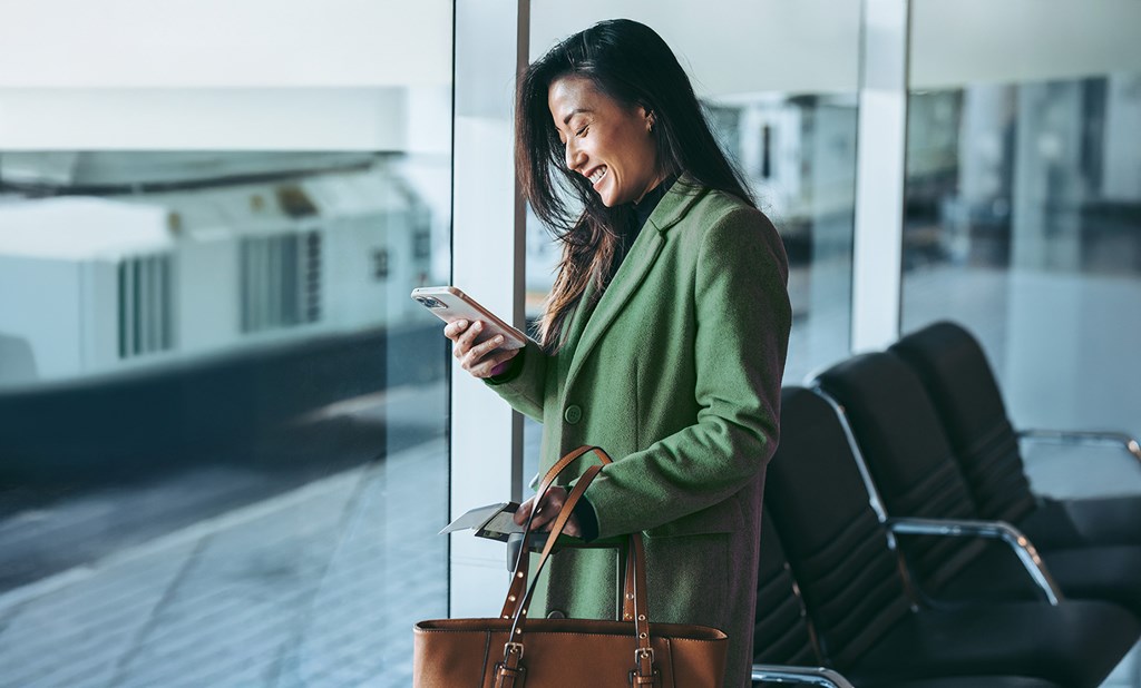 A smiling woman in a green coat at the airport looks at her mobile phone while holding travel documents.