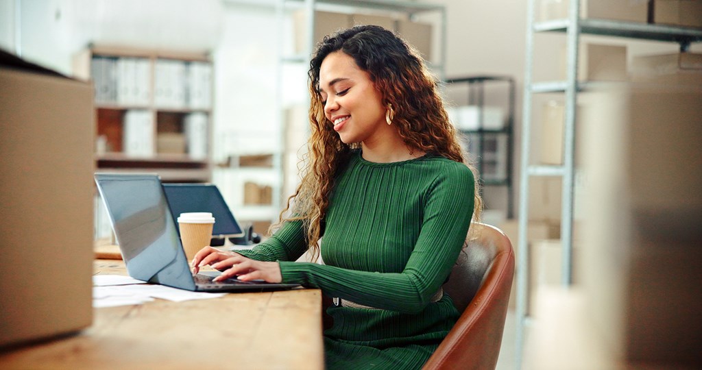 Lächelnde Frau im grünen Pullover arbeitet im Büro am Laptop, auf dem Tisch steht eine Kaffeetasse.