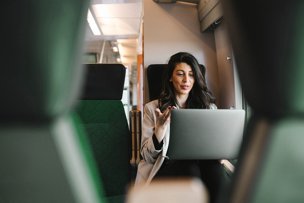 Smiling woman on a train uses a laptop, appearing to talk during a video call.