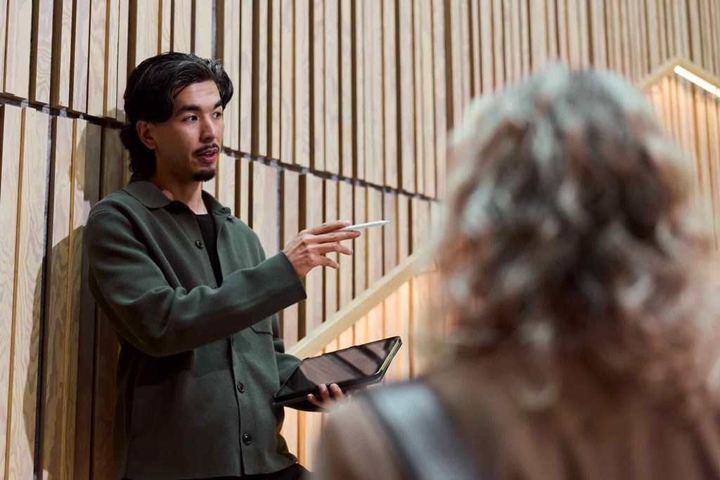 Person holding a tablet and stylus talks with another person in front of a wooden slat wall.