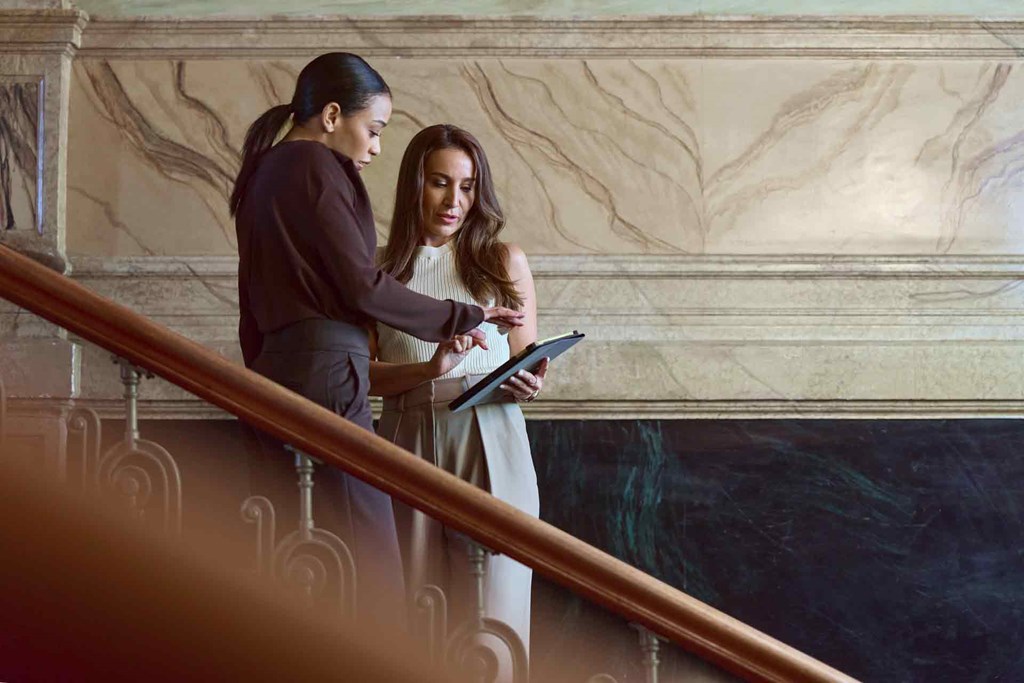 Two people stand on a staircase and look at a tablet together.