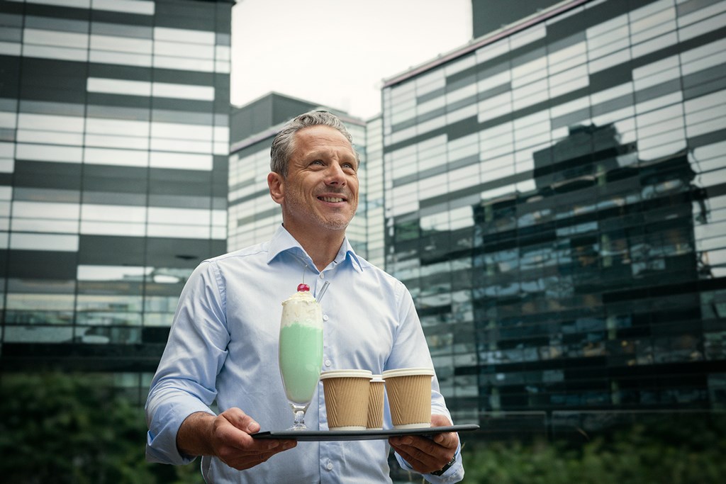 Smiling man in a light blue shirt holds a tray with two coffee cups and a green milkshake outdoors.