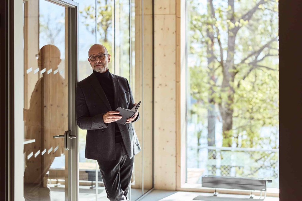 Person wearing glasses stands with an open notebook by a glass door in a bright office.