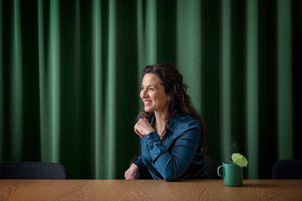 Smiling woman in a blue shirt sitting at a table with a green mug and umbrella decoration.